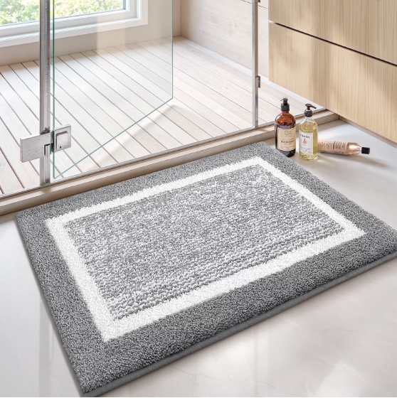 Gray bath mat with white border on a bathroom floor, next to a glass shower door.